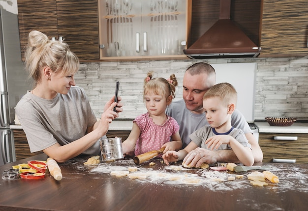 Счастливая мама фотографирует папу с двумя маленькими детьми на кухне во время приготовления мучного печенья. счастливая семья готовит вместе