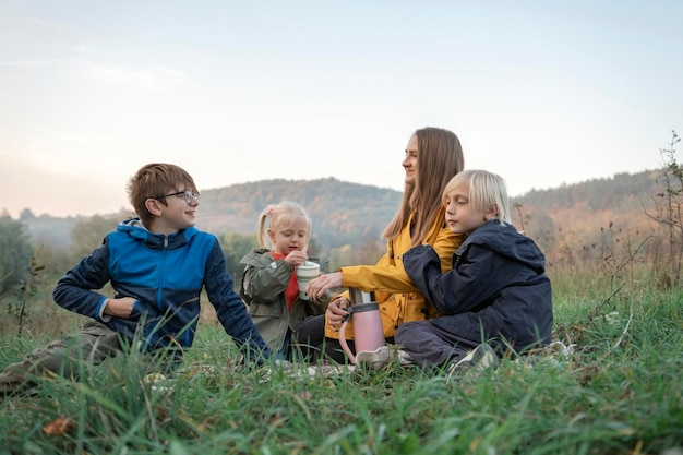 Happy mom and three kids have fun at picnic Two boys and girl with mother drink hot drink on mountains background