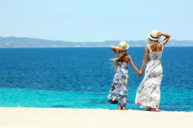 Mamma felice con la figlia in riva al mare in cappelli e abito lungo