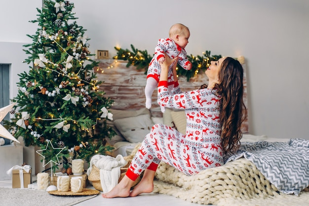 Счастливая мама с маленькой дочерью в праздничной одежде с печатными оленями и снежинками веселятся на кровати в уютной комнате с елкой и рождественскими огнями. Новый год и Рождество концепция.