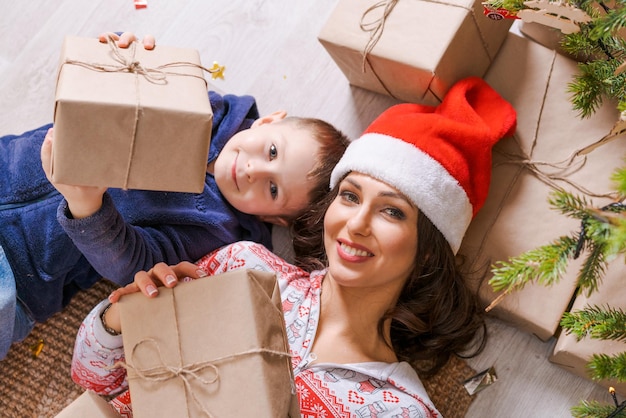 Happy mom with son in santa hat lie on floor at home under tree with gifts