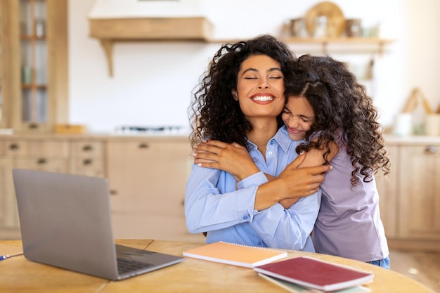 Happy mom working on laptop while her daughter embracing mother from back woman sitting at table in