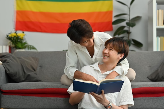 Happy moment of two young male lovers embracing each other and reading book in living room Homosexual and love concept