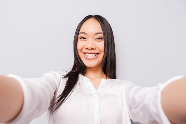 Happy moments must be saved. Cheerful young Asian woman holding camera and making selfie while standing against grey background