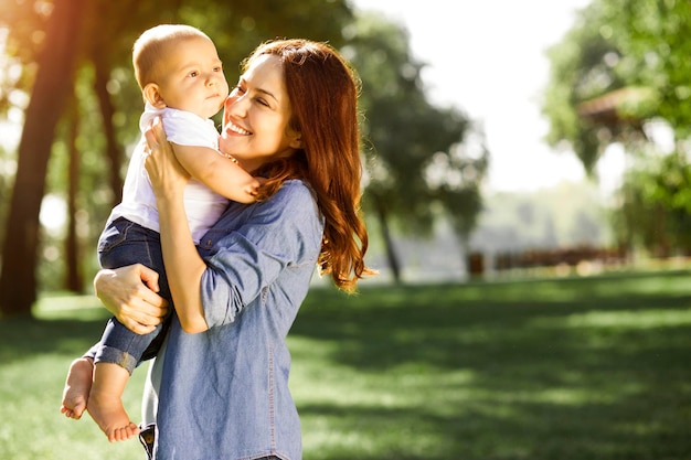 Happy mommy holding baby boy on the hands in the park with blurred background