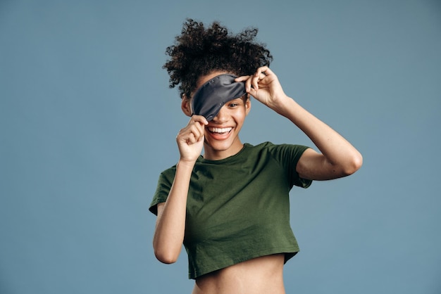Happy morning. Brunette multiracial woman wearing domestic clothes having fun with her sleeping mask and showing funny faces over the blue background