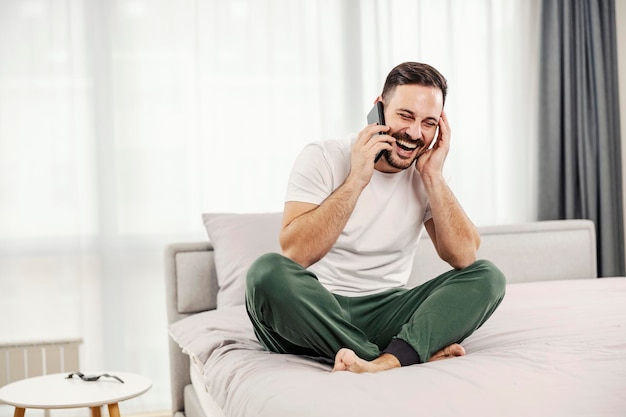 A happy morning man laughing and talking on the phone while sitting on the bed