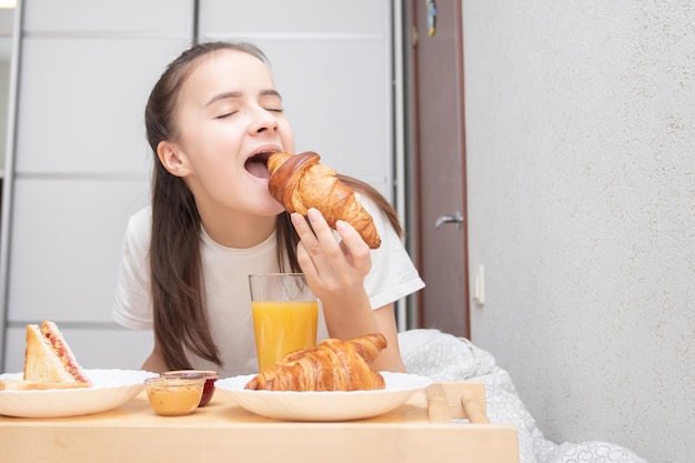 Happy morning A young woman eats a delicious Breakfast in bed  fresh croissants coffee orange juice and muesli with fruit