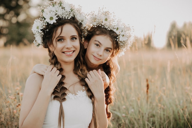 Happy mother and daughter are smiling and hugging in the field in summer in white dresses with braids and floral wreaths