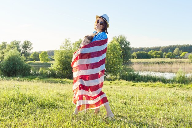 Счастливая мать и дочь девочка обнимаются под американским флагом, США, 4 июля, летний фон природы