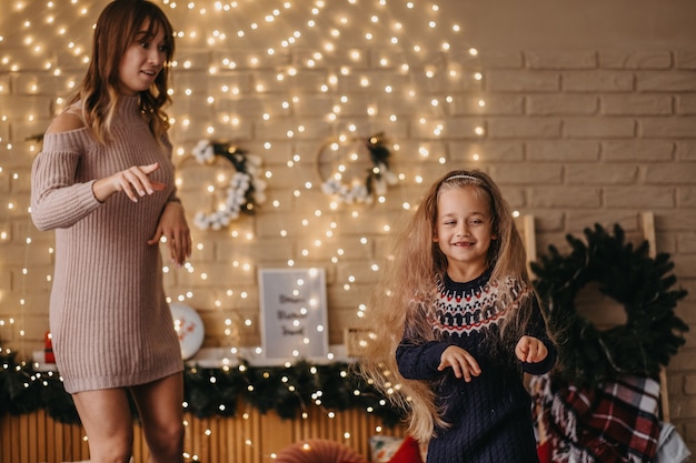 Happy mother and daughter dancing on bed, awaiting Christmas, rejoicing holidays, beautiful decoration on background