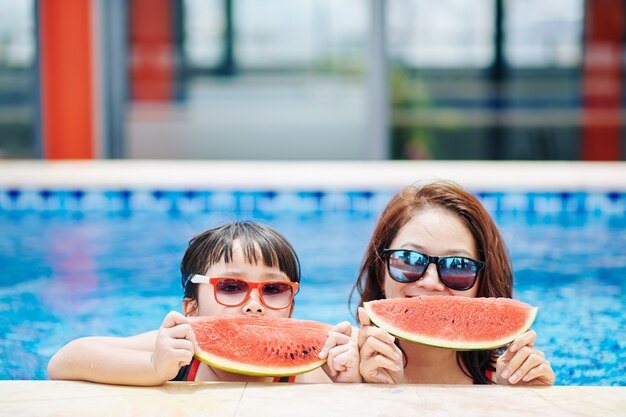 Happy mother and daughter eating watermelon after swimming in pool of spa hotel