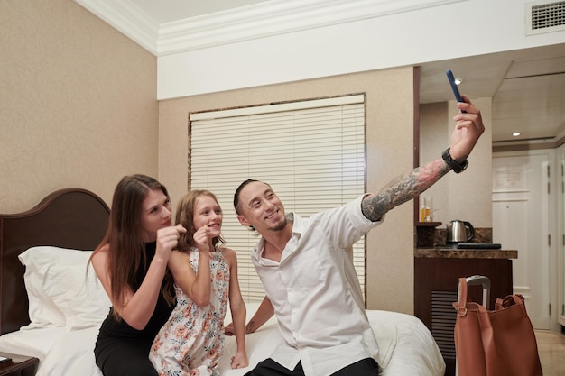 Happy mother and daughter making heart gesture with fingers when father taking selfie with them