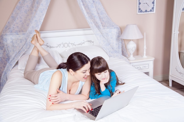 Happy mother and daughter do online shopping and pay with credit card with laptop lying on bed in bedroom. Happy family. Studio shot.