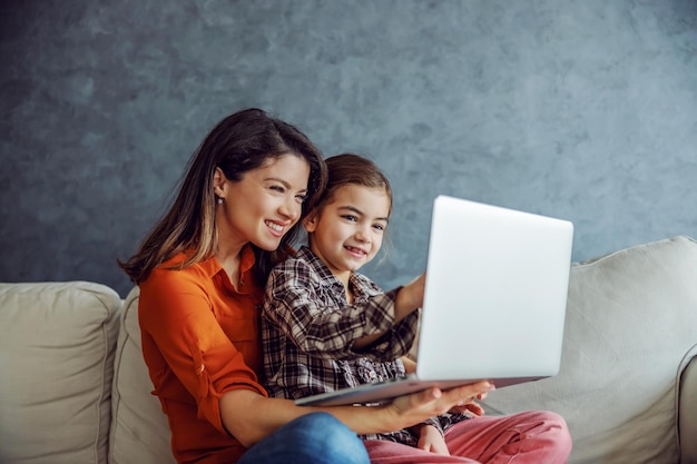 happy mother and daughter sitting on sofa and using laptop
