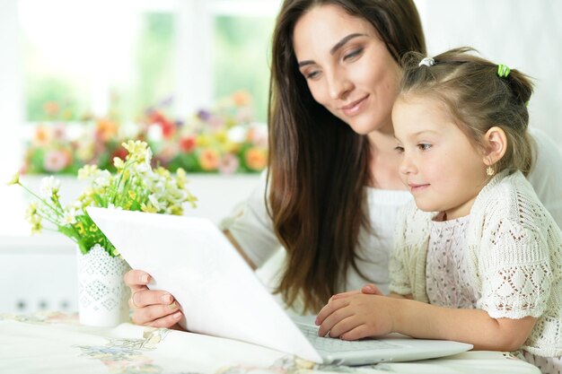 Happy mother and daughter sitting at table and using laptop