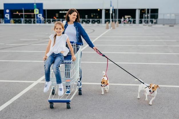 Happy mother, daughter and their pets return from shopping centre, carry cart, pose outdoor, have delighted expressions, enjoy togetherness, have friendly relationships, buy something. Family concept.