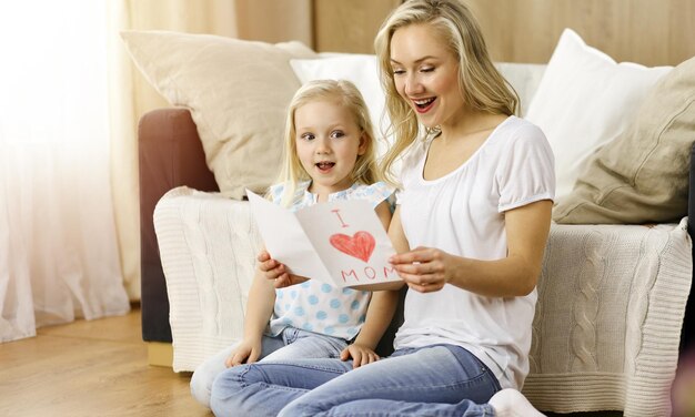 Happy mother day in sunny flat. Child daughter congratulates mom and gives her postcard with heart drawing. Family concept.