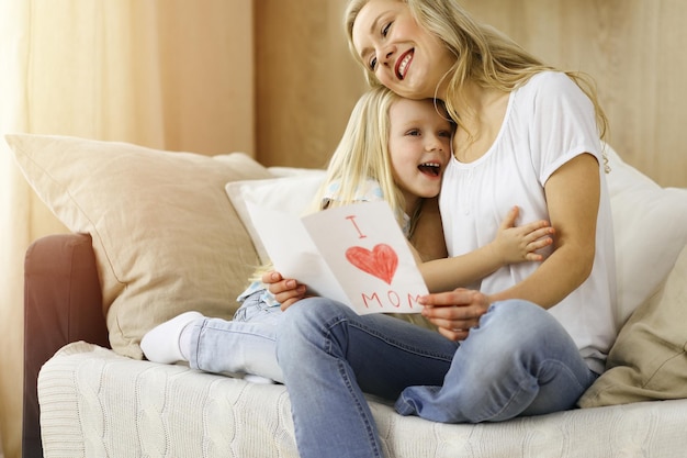 Happy mother day in sunny flat. Child daughter congratulates mom and gives her postcard with heart drawing. Family concept.