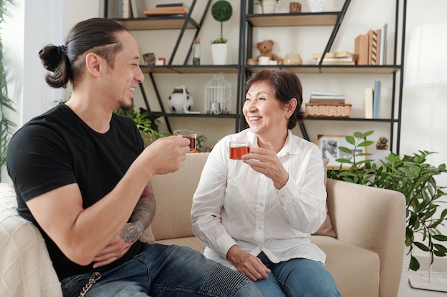 幸せな母親がお茶を飲み、リビングルームで息子とソファで会話中に笑う