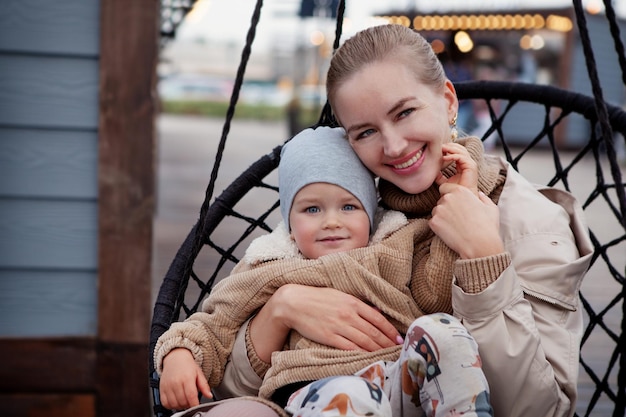사진 해피 어머니는 웃으면서 카메라를 보고 있는 아이를 포용합니다. 아름다운 젊은 엄마와 아기가 껴안고 즐거운 시간을 보내세요. 니트 베이지색 스웨터, 재킷, 비니를 입은 모델. 라이프 스타일 초상화
