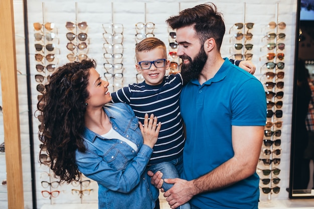 Happy mother and father choosing eyeglasses frame for their son in optical store.