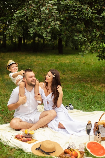 Happy mother, father and their cute little son s having picnic at summer park. Child sitting on his father shoulders. Family and leisure concept