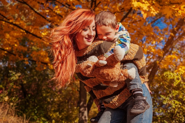 Happy mother and her little son walking and having fun in autumn forest. Kid riding on mother's back