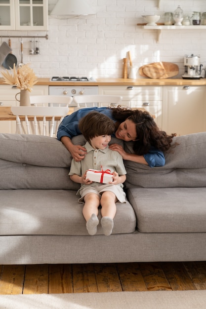 L'abbraccio felice della madre ha sorpreso il ragazzo di compleanno prescolare allegro con la scatola regalo avvolta seduto sul divano