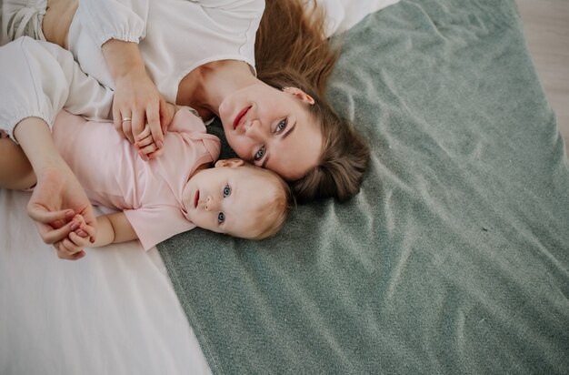 A happy mother is lying with her baby daughter on the bed in the room. Top view