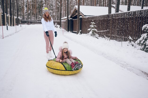 Happy mother and little cute girl in pink warm outwear walking having fun rides inflatable snow tube