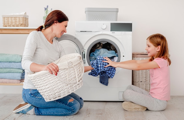 Madre felice e piccola figlia che lavano i vestiti facendo uso della macchina nella stanza leggera