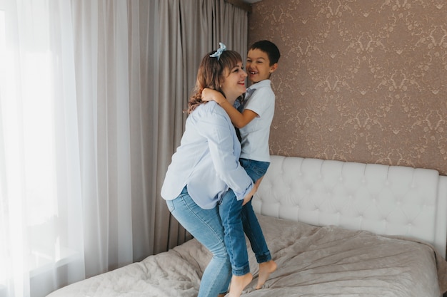 Happy mother and little son are jumping on the bed, holding hands, laughing mom is playing a game with the child in the bedroom.