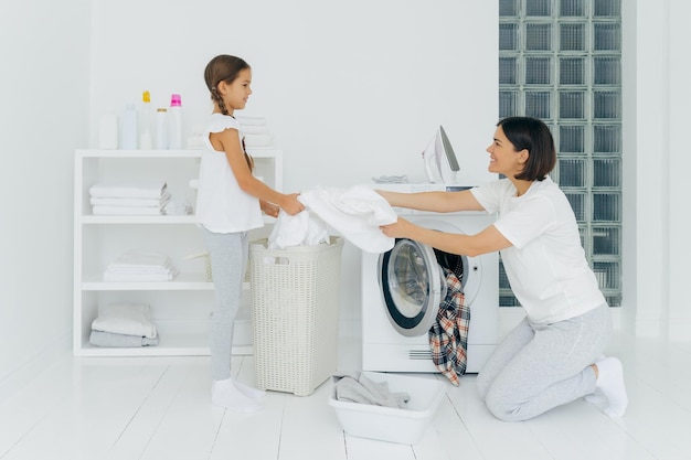 Happy mother loads clothes in washing machine little girl helps gives white linen from basket do housework together dressed in casual clothing Daughter offers help to mum clean clothes