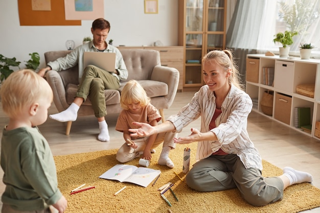 Madre felice che gioca sul pavimento insieme ai suoi figli mentre il padre lavora al computer portatile seduto sul divano di casa