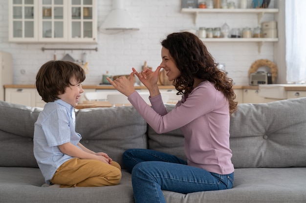 Happy mother playing with her child on couch at home, makes funny grimace