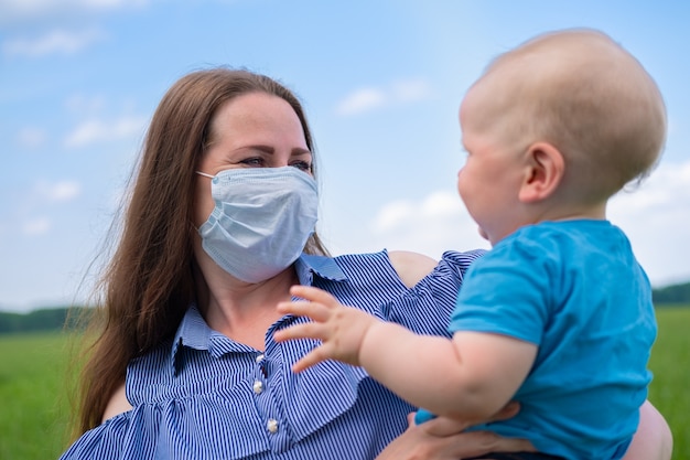 La madre felice nella mascherina medica protettiva tiene il suo piccolo figlio sulle braccia in natura