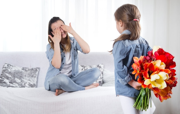 母の日おめでとう。チューリップの大きな花束を持つ小さな甘い娘が母親を祝福します。リビングのインテリアは、幸せな家庭生活のコンセプト