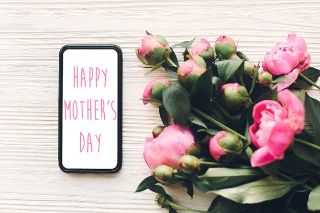 Foto happy mother's day tekst op telefoonscherm en roze pioenrozen op rustieke witte houten achtergrond in lichte bloemen wenskaart concept plat lag moederdag tedere lente afbeelding
