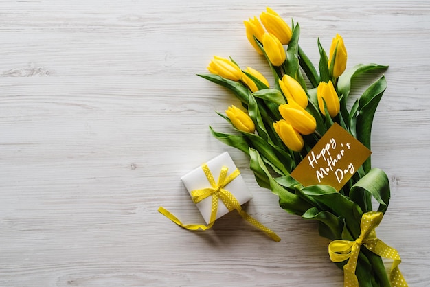 Happy mother's day text sign on craft postcard Bouquet of tulips with gift box on white wooden background Holiday greeting card for Mother's Day Top view flat lay