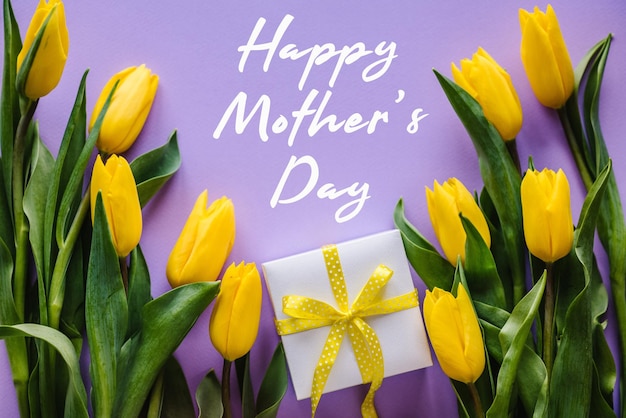 Happy mother's day Text sign with yellow tulips and gift box on violet background Floral greeting card concept Holiday greeting card for Mother's Day Top view flat lay