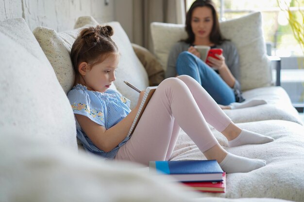 Happy mother smilling daughter sitting on sofa enjoying creative activity, drawing pen pictures in albums, mother and daughter spend free time together.