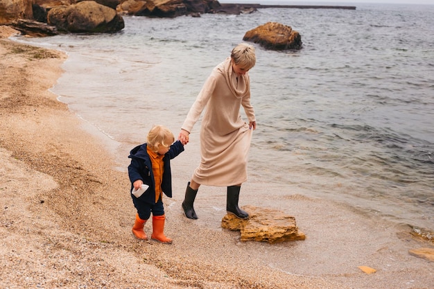 Happy mother and son playing with paper boat on beach in autumn time