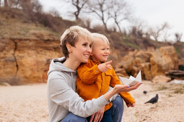 Happy mother and son playing with paper boat near sea on seashore in autumn time