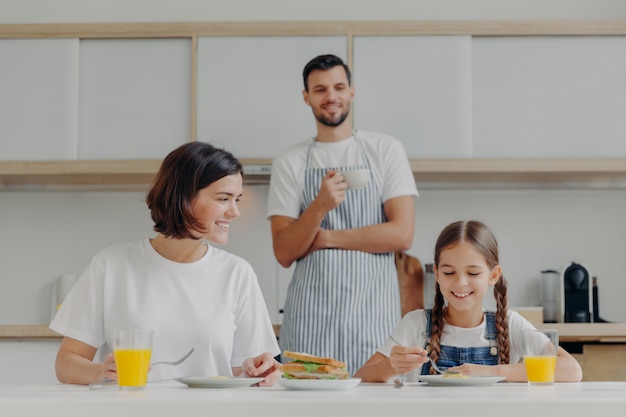 Happy mother talks to daughter while have breakfast. Father stands behind, prepared delicious dish for family.