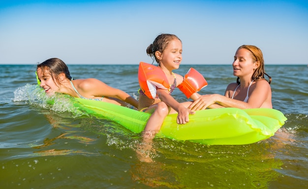 Счастливая мама и две маленькие позитивные дочки купаются и плавают в море на надувном матрасе