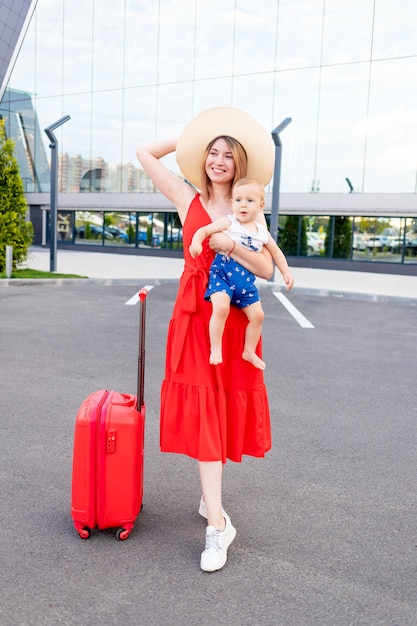 A happy mother with a baby son at the airport with a red suitcase goes on a trip or vacation in the summer