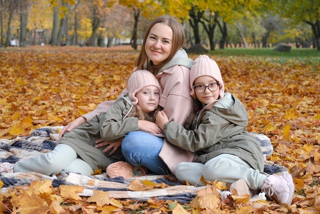 双子の娘たちと幸せな母親が秋の公園で毛布に座って時間を過ごす