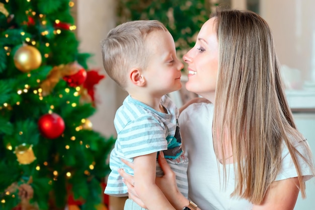 ゆるい髪の幸せな母親は、クリスマスツリーの背景に彼女の幼い息子を抱きしめます休日を待っています