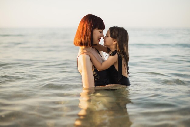 Happy mother with red hair holding daughter in a black swimming suit in the sea